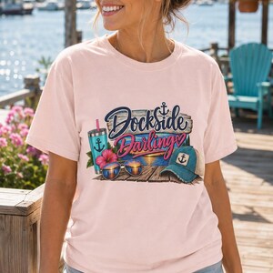 May include: A light pink short-sleeve t-shirt featuring a graphic print of a wooden dock with the text Dockside Darlings. The design includes a teal tumbler with an anchor, a pink hibiscus flower, sunglasses reflecting a sunset, and a baseball cap.