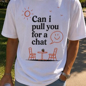以下が含まれることがあります： 黒とオレンジのグラフィックプリントが施された白い半袖Tシャツ。デザインには「Can i pull you for a chat」というテキストに加え、太陽、鳥、笑顔の吹き出し、小さなテーブルを囲む2脚の椅子が描かれています。