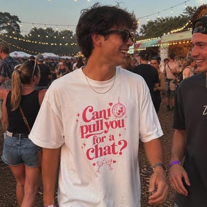 May include: A white short-sleeve t-shirt featuring pink graphic text that reads "Can I pull you for a chat?" accompanied by a disco ball, small stars, hearts, and two clinking cocktail glasses printed on the centre of the chest.