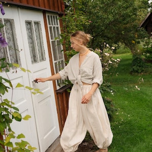 Könnte beinhalten: Ein beiges, langärmeliges Hemd mit Knopfleiste und geknotetem Saum sowie eine dazu passende, weit geschnittene beigefarbene Hose mit elastischen Bündchen an den Knöcheln. Das Outfit wird durch braune Sandalen mit zwei Riemen und eine orangefarbene Perlenkette ergänzt.