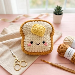 May include: A crocheted plush toy shaped like a slice of toast with a golden yellow butter square on top. The toast has a cream-colored center, a dark brown crust, black bead eyes, a small stitched smile, and pink embroidered cheeks.