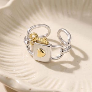 May include: A silver-toned open band ring featuring a central rectangular charm shaped like a padlock with a small gold-toned heart accent. A tiny gold-toned key charm is attached to the top of the padlock, and the band consists of linked silver-toned loops.