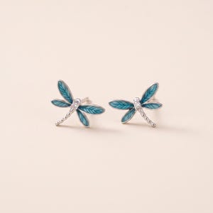 May include: A pair of silver stud earrings shaped like dragonflies. Each earring features four wings filled with teal enamel and a small clear stone set in the centre of the body. The dragonfly bodies have a segmented, textured silver design.
