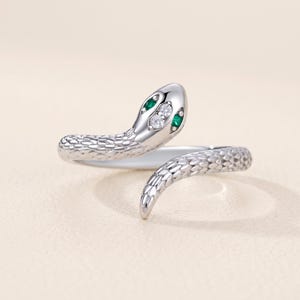 Puede incluir: Un anillo de serpiente en tono plateado con un patrón de escamas texturizado a lo largo de la banda. La cabeza de la serpiente está adornada con dos pequeñas piedras redondas transparentes y dos piedras verdes en forma de marquesa como ojos.
