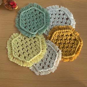 May include: A set of five hexagonal macrame coasters featuring a knotted cord pattern with fringed edges. The coasters are coloured in sage green, mustard yellow, light yellow, and two shades of off-white, arranged in an overlapping cluster.