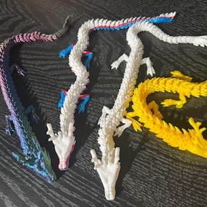 Può includere: Quattro statuette di drago articolate con corpi segmentati. Una è viola e blu iridescente, una è bianca con accenti blu, una è bianco solido e una è giallo solido. Ogni drago presenta squame dettagliate, quattro zampe e corna.