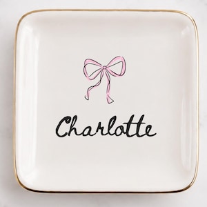 Peut inclure: Un vide-poche carré en céramique blanche avec un bord doré. Le centre présente une illustration de nœud rose dessinée à la main au-dessus du prénom Charlotte écrit en écriture cursive noire. Le plat a une finition lisse et brillante.