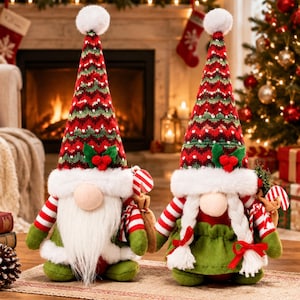 May include: Two plush Christmas gnomes with tall, pointed hats featuring red, green, and white chevron patterns. One gnome has a long white beard, while the other has two white braided pigtails. Both wear white faux fur trim.