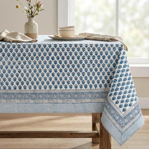 May include: A rectangular tablecloth featuring a repeating blue floral block print pattern on a white background, accented by a wide border with intricate floral motifs and thin blue and white stripes along the bottom edge of the fabric.