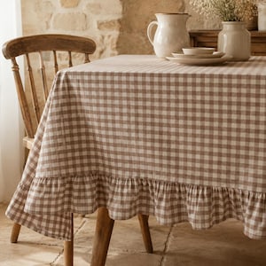 May include: A rectangular tablecloth featuring a brown and white gingham check pattern with a ruffled edge. The fabric drapes over a wooden table, which is accompanied by a wooden chair. A ceramic pitcher and stacked plates sit on the table.
