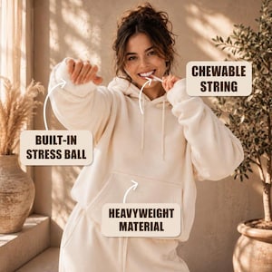 May include: A cream-colored hooded sweatshirt featuring a built-in stress ball in the sleeve, a chewable string at the neckline, and a heavyweight material construction. The garment includes text labels pointing to these specific functional features.