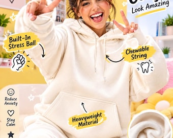 Puede incluir: Una sudadera con capucha de color crema que cuenta con un bolsillo tipo canguro, capucha con cordón y características sensoriales integradas, incluyendo una pelota antiestrés y un cordón masticable. El tejido es de forro polar grueso y suave.