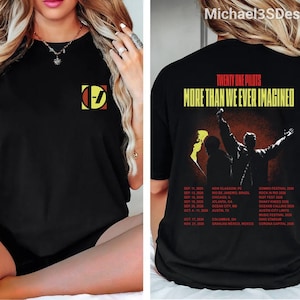 Op de afbeelding: Een zwart t-shirt met een klein Twenty One Pilots-logo op de linkerborst. De achterkant toont de tekst "TWENTY ONE PILOTS" en "MORE THAN WE EVER IMAGINED" boven een afbeelding van een artiest en een lijst met tourdata voor 2026.