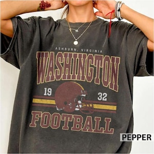 May include: A charcoal grey short-sleeve t-shirt featuring a distressed graphic print. The text reads ASHBURN, VIRGINIA, WASHINGTON, and FOOTBALL, with the numbers 19 and 32 flanking a maroon football helmet centred above a yellow stripe.