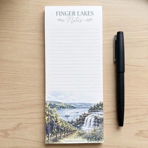 Könnte beinhalten: Ein rechteckiger Notizblock mit horizontalen Linien und dem Text FINGER LAKES Notes am oberen Rand. Der untere Teil zeigt eine Aquarellillustration eines Weinbergs, eines Sees mit Segelbooten und eines Wasserfalls. Ein mattschwarzer Stift liegt daneben.