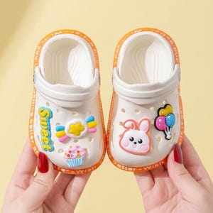 May include: A pair of white slip-on clogs with orange trim featuring the text "Sweet" and "Wish" along the edges. The shoes are decorated with colorful rubber charms including a star candy, a cupcake, a white rabbit face, and balloons.
