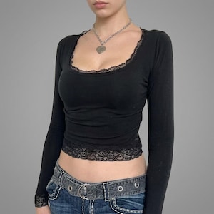 May include: A black long-sleeve top featuring a scoop neckline trimmed with black lace, matching lace detailing at the cuffs and hem, and a silver-toned chain necklace with a heart-shaped pendant, paired with a dark denim belt with metal hardware.