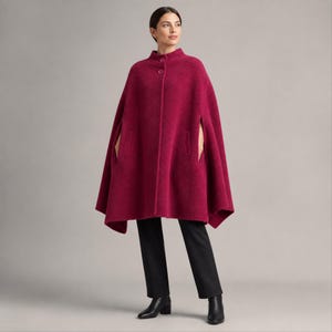 Peut inclure: Une cape magenta en mélange de laine avec un col montant, deux boutons visibles au niveau de l'encolure et des fentes latérales pour les bras. Le vêtement présente une silhouette structurée et évasée tombant sous les hanches, avec un pantalon noir.