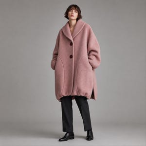 May include: A dusty pink oversized wool coat featuring a wide shawl collar, dropped shoulders, and two large dark brown buttons down the front. The coat has a rounded hemline and is paired with straight-leg charcoal gray trousers and black leather shoes.