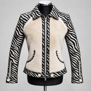 Könnte beinhalten: Eine Jacke mit schwarz-weißem Zebramuster an Ärmeln, Kragen und Besatz, kontrastiert durch cremefarbene Kunstfell-Einsätze auf der Vorderseite. Das Kleidungsstück verfügt über einen zentralen silbernen Reißverschluss, zwei Seitentaschen und weiße Knöpfe.