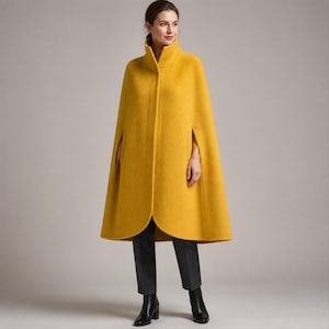 Peut inclure: Un manteau cape en laine jaune moutarde doté d'un col montant, d'une fermeture boutonnée dissimulée sur le devant et de fentes pour les bras sur les côtés. Le vêtement présente une silhouette trapèze structurée tombant sous les hanches.