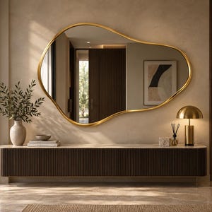 Puede incluir: Un gran espejo de pared con una forma orgánica e irregular similar a una ameba, con un marco fino de metal en tono dorado pulido. La superficie reflectante muestra el interior de una habitación con una consola flotante de listones.