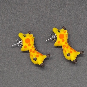 Pode incluir: Um par de brincos de tarracha em forma de girafas amarelas com manchas laranja, cascos castanho-escuros e pequenos chifres. Os brincos possuem pinos de metal prateado e fechos de borboleta, desenhados para parecer que a girafa morde.