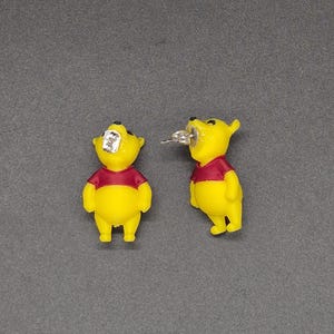 Pode incluir: Um par de brincos de pino com o formato de um urso amarelo vestindo uma camisa vermelha. Um brinco é mostrado de frente, enquanto o outro está virado de lado, revelando o pino de metal prateado e a tarracha de borboleta.