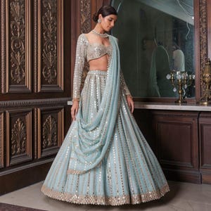 May include: A light blue lehenga set featuring a long-sleeved blouse and a floor-length skirt, both heavily embellished with reflective circular mirror work and intricate gold embroidery. A matching sheer dupatta drapes over the shoulder.