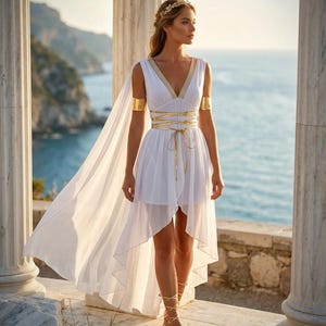 Pode incluir: Um vestido branco sem mangas com bainha assimétrica, com acabamento dourado ao longo do decote em V e detalhe de cordões dourados na cintura. O vestido inclui uma capa longa e fluida presa aos ombros, braceletes dourados e sandálias romanas.