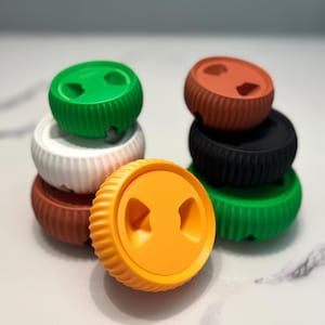 Puede incluir: Una colección de organizadores de cables circulares y acanalados en varios colores, incluyendo verde, blanco, marrón, naranja y negro. Cada organizador presenta una parte superior plana con dos recortes triangulares y un perfil lateral estriado.