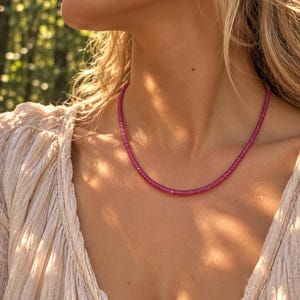 Puede incluir: Un collar compuesto por pequeñas cuentas redondas y facetadas de color magenta. Las cuentas son uniformes en tamaño y forma, ensartadas para formar una hebra circular continua que descansa sobre el cuello. La superficie refleja la luz.