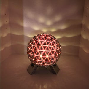 May include: A spherical lamp featuring a geometric triangular grid pattern that emits a warm, glowing light. The orb is supported by a dark, angular base, casting soft, wave-like shadows against the surrounding enclosure.