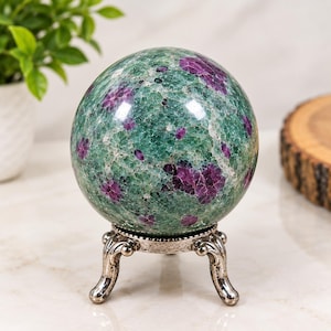 May include: A polished spherical ruby in zoisite gemstone resting on a decorative silver-toned metal tripod stand. The stone features a mottled texture with deep magenta-pink ruby inclusions embedded within a vibrant green zoisite matrix.