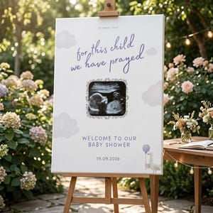 Op de afbeelding: Een wit rechthoekig bord op een houten ezel met de tekst "for this child we have prayed", "1st Samuel 1:27", "WELCOME TO OUR BABY SHOWER" en "19.09.2026". Het bevat een centrale echofoto, grijze wolken en een kleine vogel met een ballon.