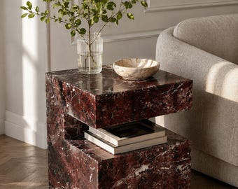 May include: A C-shaped side table crafted from deep red marble with prominent white veining. The table features a hollow middle section for storage, currently holding two stacked books, and a flat top surface supporting a glass vase and bowl.