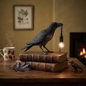 May include: A black raven-shaped lamp standing on two stacked vintage leather-bound books. The raven holds a black cord with a glowing round light bulb in its beak. A small bronze-colored bird figurine sits nearby on the wooden surface.