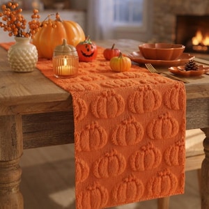 Könnte beinhalten: Ein orangefarbener Tischläufer mit einem sich wiederholenden, erhabenen Kürbismuster. Der Läufer liegt auf einem Holztisch, dekoriert mit einer weißen, strukturierten Vase mit orangefarbenen Beeren, einem großen Kürbis, einer Kerze und kleinen Kürbissen.