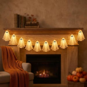 Könnte beinhalten: Eine Lichterkette mit zehn geisterförmigen Leuchten, die mit einem weißen, spitzenartigen Stoff überzogen sind und jeweils zwei dunkle, ovale Augen aufweisen. Die Geister sind durch einen dünnen Draht verbunden und strahlen ein warmes Licht aus.