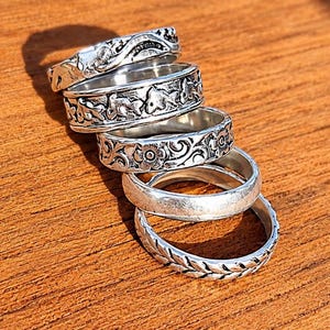 Op de afbeelding: Een stapel van vijf zilverkleurige metalen ringen met diverse ontwerpen, waaronder bloemmotieven, bladpatronen en getextureerde oppervlakken. De ringen zijn in een verticale lijn gerangschikt en tonen verschillende breedtes en reliëfdetails.