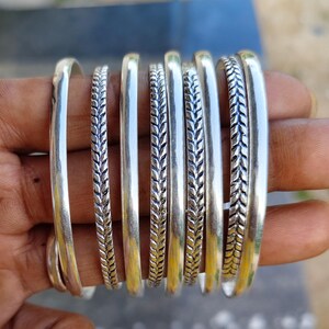 May include: A collection of eight silver-toned bangles arranged side-by-side. The set features alternating designs, including smooth, polished bands and textured bands with a repeating leaf-like or braided pattern along the outer surface.