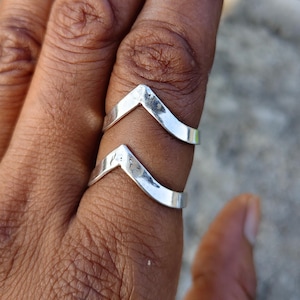 May include: Two silver-coloured rings with a chevron or V-shaped design. Each ring features a smooth, polished metallic surface that forms a pointed angle at the centre, worn stacked on a finger to create a layered appearance on the hand.