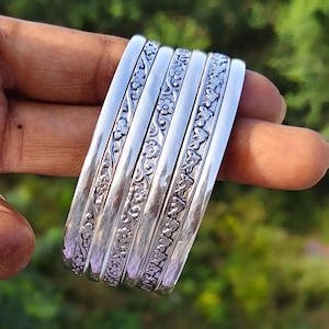Könnte beinhalten: Ein Set aus fünf silberfarbenen Armreifen mit abwechselnden Bändern aus glattem, poliertem Metall und vertieften Abschnitten mit einem detaillierten Blumenrankenmuster. Die Armreifen sind nebeneinander angeordnet und zeigen ein einheitliches Finish.
