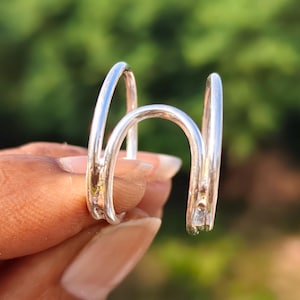 May include: A silver-coloured ear cuff featuring a double-banded design with a smooth, polished finish. The piece consists of two parallel curved metal wires connected by a central bridge, creating an open, structural shape for ear wear.