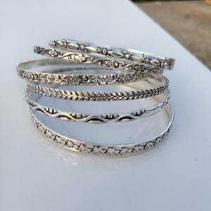 May include: A stack of six silver-toned metal bangles, each featuring a distinct embossed pattern. The designs include floral motifs, braided textures, and wavy lines. These circular bracelets are arranged in a slightly overlapping vertical pile.
