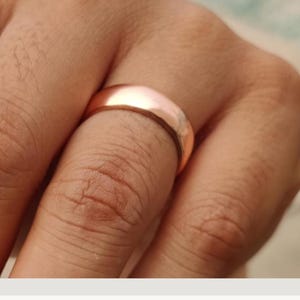 Può includere: Un anello a fascia in oro rosa liscio e lucido con profilo arrotondato. La superficie metallica ha una finitura riflettente che cattura la luce, mostrando un design circolare semplice e continuo senza incisioni o elementi decorativi.