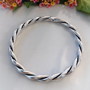Può includere: Un bracciale rigido circolare realizzato in metallo color argento lucido. Il design presenta un motivo a corda intrecciata continua con ossidazione scura e incassata nelle scanalature tra i fili, creando un aspetto strutturato ad alto contrasto.
