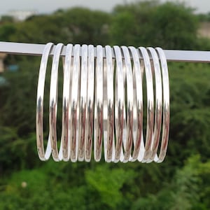 Pode incluir: Um conjunto de doze pulseiras finas de cor prateada, polidas e dispostas em fila. Cada pulseira apresenta uma superfície lisa e arredondada com um acabamento metálico refletor. As pulseiras estão alinhadas verticalmente numa barra.