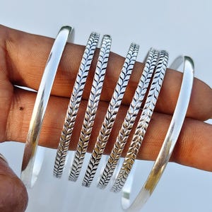 May include: A set of six silver-toned bangles. Two bangles feature a smooth, polished surface, while four bangles display a detailed, repeating leaf or wheat-like pattern etched along the entire outer circumference of each individual piece.