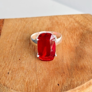 May include: A silver ring featuring a large, rectangular, faceted red gemstone set in a four-prong mounting. The stone has a deep crimson hue with visible light reflections on its polished surface, and the band is a smooth, polished silver metal.